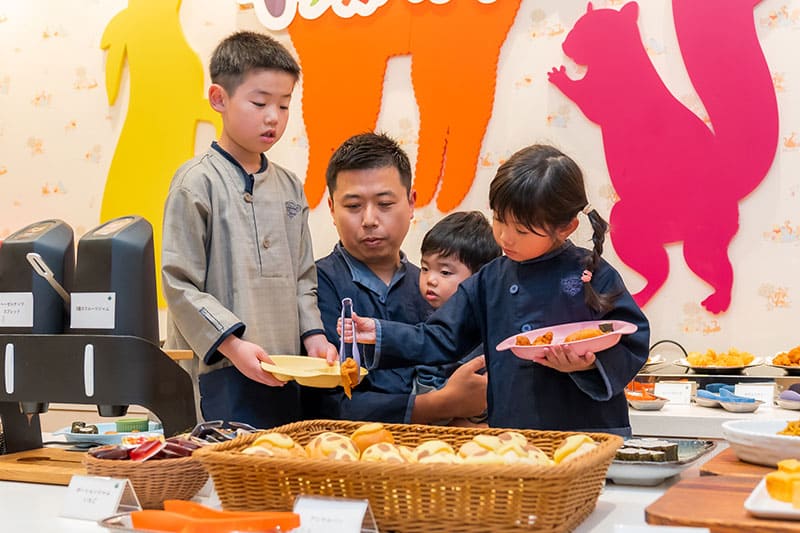 小さい女の子がトングで料理を取ろうとしている写真