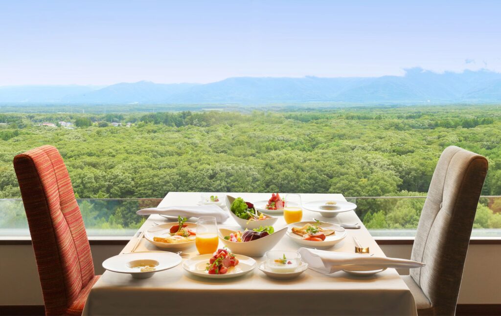 窓の向こうに山並みが見える2人席。料理が机上に並ぶ写真