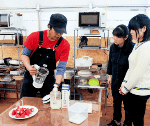 たくみの里　いちごの家のハウス内で、男性スタッフが女性客2人を前に、いちごのスムージー作りの説明をしている写真